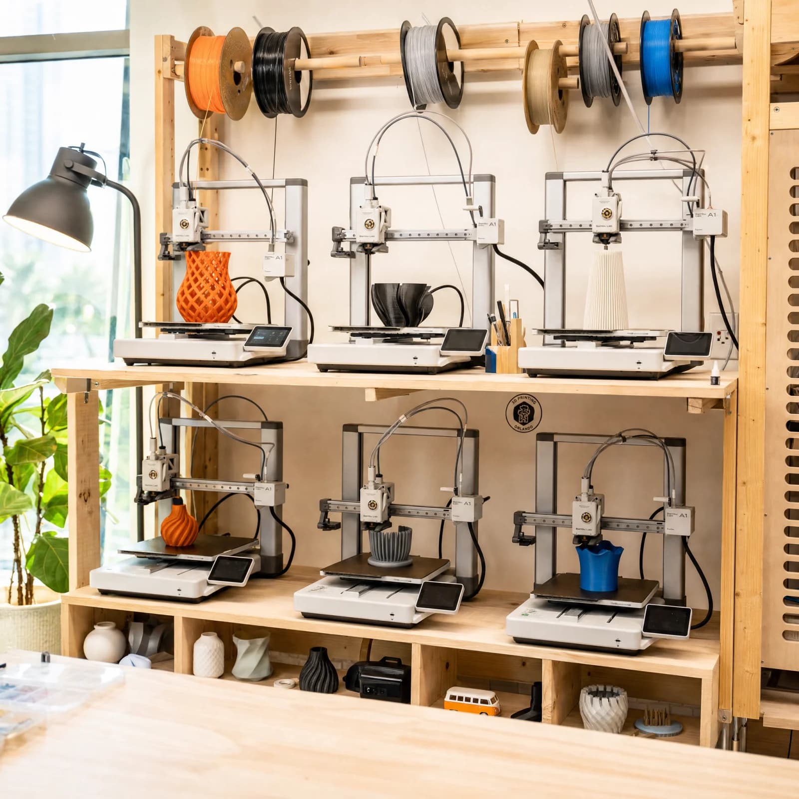 The AM3D Orlando studio — six Bambu printers actively printing on wood shelving with filament spools above